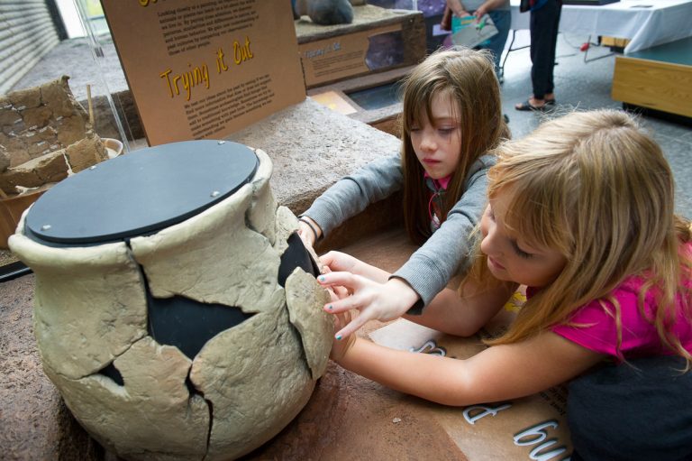 Two young girls lean over a display and fit pieces of a broken pot back together.