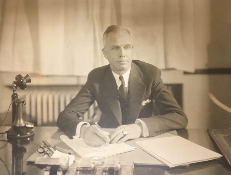 Ewert, in a suit, sits at his desk, holding a pen as if he's about to begin writing. Papers and a telephone are visible on his desk. Sepia photograph.