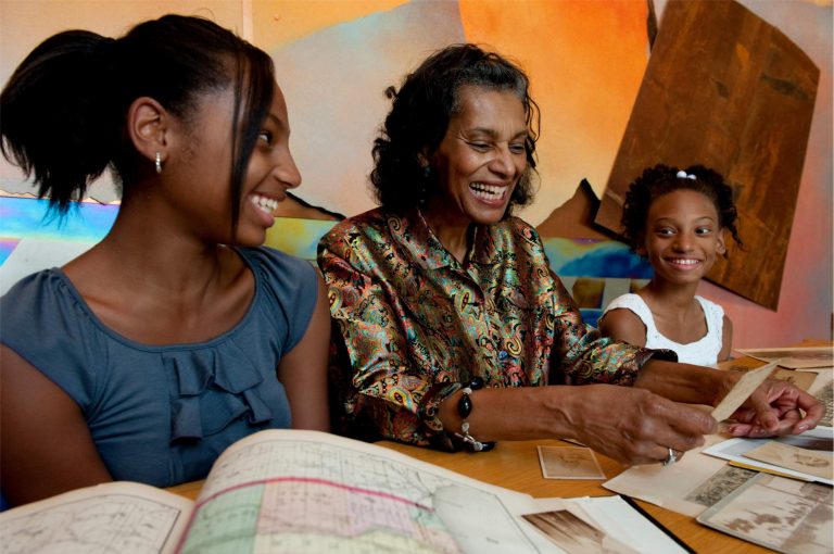 Adult woman and two young girls smile and laugh while paging through historic documents laid out on a wood table.