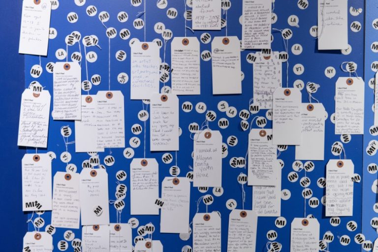 Blue wall with dozens of white tags with hang written notes hanging from it. Stickers with state abbreviations also on wall.