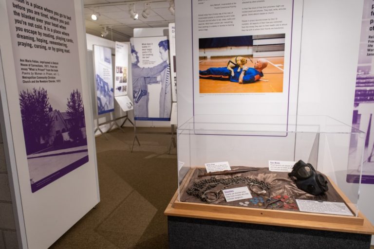 Standing exhibit panels in a gallery. A glass case containing shackles and a gas mask visible in front of panels.