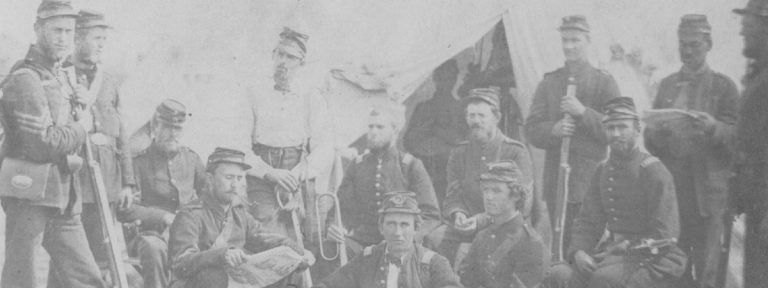 Men of the Sixth Michigan Infantry cluster together. Some are standing with rifles, some are sitting with instruments.