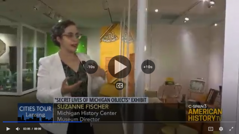 Video still; Suzanne Fischer. Overlaid title reads: "Secret lives of Michigan Objects" exhibit stands in a museum exhibit.