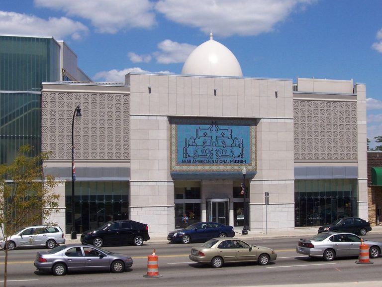 Exterior - light stone building with tall windows and a large blue sign reading "Arab American National Museum"