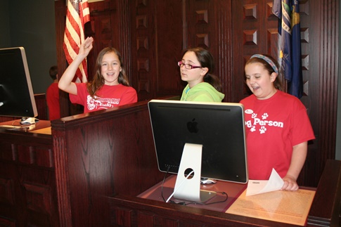 Three middle school-aged students stand behind an imposing wood desk with a computer monitor on it.