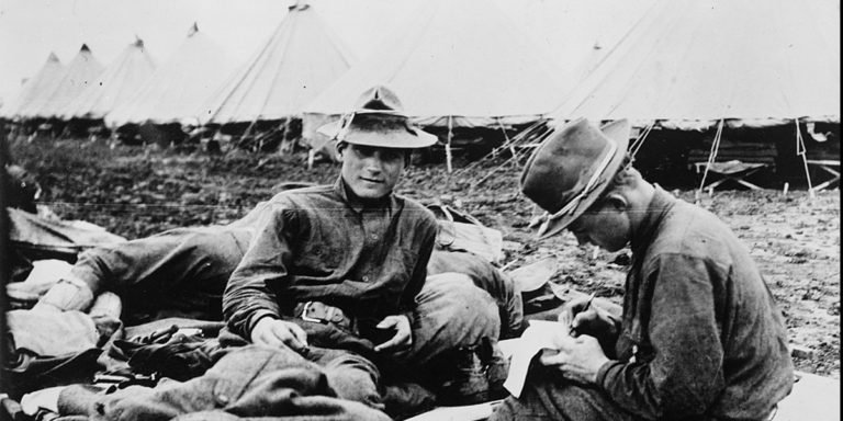 Photograph shows three soldiers sitting on the ground with bedding, blankets, and rifles. One soldier is writing a letter and smoking, another looks toward the camera while behind them the other soldier is sleeping. Several white bell tents are in the background.